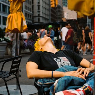 Person with yellow hair leaning back in a chair on a busy city sidewalk, eyes closed.