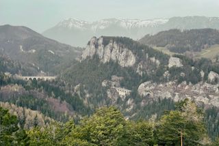 In the foreground a densly forrested rock face with train tracks leading through a tunnel and across multiple viaducts and in the background are taller mountains that are still covered in snow