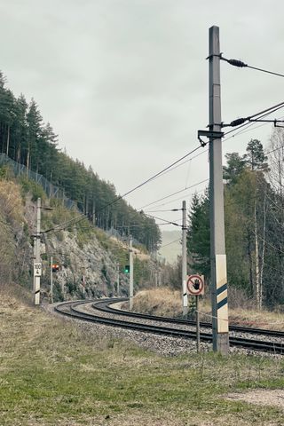 Train tracks leading through a mountains area, on the left is a rock face with trees on top of it