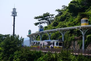 A train with yellow and blue cars travels along an elevated track with a larger yellow train traveling on a lower level, next to a lush green hillside, with people on a nearby walkway and a communication tower in the background.