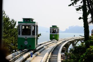 Two green monorail cars on an elevated track overlooking the ocean and a distant city, with trees in the foreground.