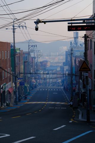 A street with many overhead power lines and buildings on both sides leads into a hazy distance, with mountains in the background.