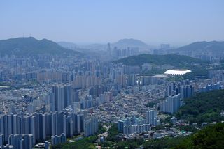 An aerial view of a sprawling city with numerous high-rise buildings and a large stadium, surrounded by green hills under a hazy sky.