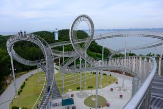 An intricate, looping silver pedestrian bridge with people walking on it, overlooking a green island and the ocean with a lighthouse in the distance.