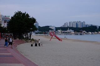 A wide sandy beach with a red life guard chair, a busker, and people walking on a promenade, with a city skyline and mountains in the background under a twilight sky.