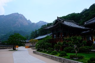 A traditional Korean temple with ornate roofs and lush gardens, set against a backdrop of rugged mountains under a cloudy sky.