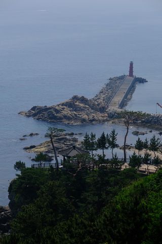 An aerial view of a red lighthouse at the end of a pier, extending from a rocky coastline into the calm blue sea, with a traditional gazebo and pine trees on a green cliff in the foreground.