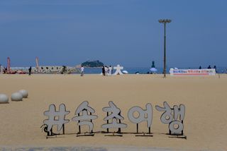 A wide sandy beach with a large sign featuring Korean characters in the foreground, people walking, and a distant island with a lighthouse under a clear blue sky.
