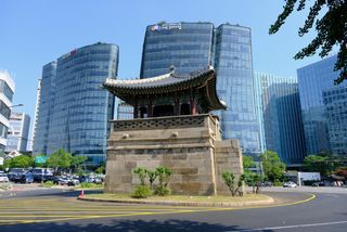 A traditional Korean pavilion stands in the middle of a traffic circle, surrounded by modern glass skyscrapers under a clear blue sky.