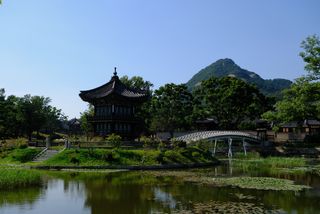 A traditional Korean pavilion sits on an island in a calm lake with a white arched bridge, surrounded by lush trees and a mountain in the background under a clear sky.