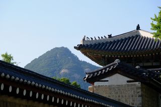 Traditional Korean temple rooftops with ornate decorations are visible in the foreground, with a lush green mountain under a clear sky in the background.