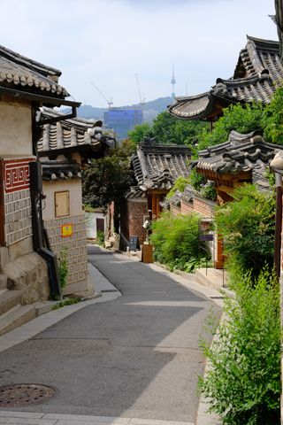 A narrow, winding street lined with traditional Korean houses with tiled roofs and green foliage, leading towards a modern city skyline with a tower in the distance.