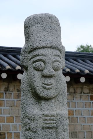 A close-up of a smiling stone statue with a tall hat, set against a traditional Korean wall and tiled roof.