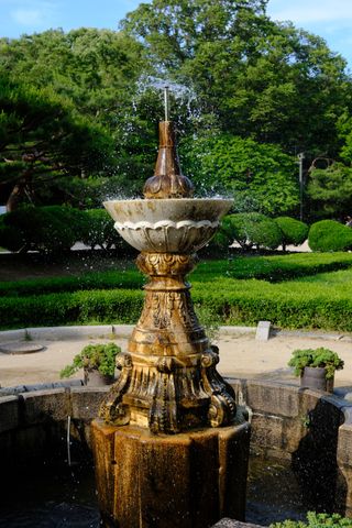 An ornate stone fountain sprays water in a lush green garden with trees in the background.