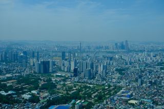 An aerial view of a sprawling city with numerous high-rise buildings, a river, and distant mountains under a hazy sky.
