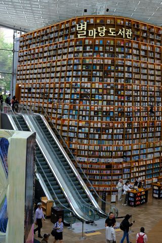 A large, multi-story library with towering bookshelves filled with books, an escalator, and people browsing and walking below.