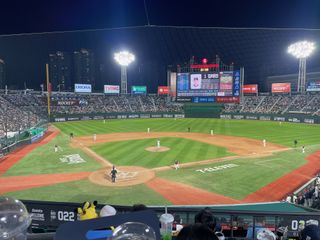 A wide shot of a baseball stadium at night, with players on the field, a large scoreboard displaying game information, and spectators in the stands, with city buildings visible in the background.