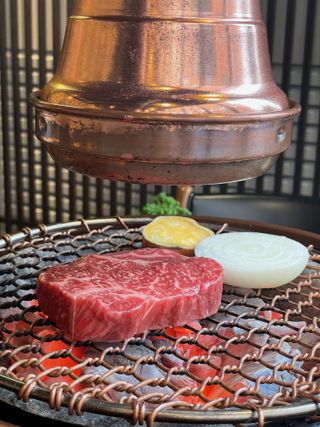 A raw cut of marbled beef, a slice of onion, and a small dish on a grill with hot coals, under a copper exhaust hood.