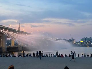 People gathered by a river at dusk, watching a bridge with a water fountain display under a colorful sky.