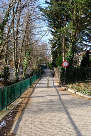 Footpath and on the right side is a fence and some trees and on the left side is a green railing separating the footpath from an embankment; the weather is sunny and there are some tiny specks of snow