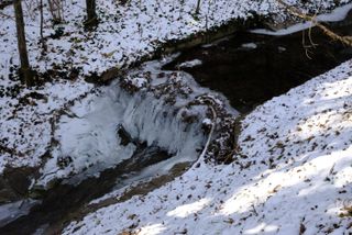 A tiny stream running from the right to the left and there is a tiny waterfall, like 50 cm high, and some parts of the waterfall are frozen; snow lays on both sides of the stream