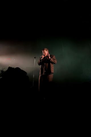 A dark, moody concert photo of Kae Tempest in a brown suit jacket, singing into a microphone with one hand raised near their ear monitor, backlit by a hazy green glow with silhouetted audience heads in the foreground.