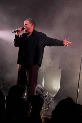 Kae Tempest sings into a handheld microphone on a smoky, dimly lit stage, wearing a black button-up shirt and dark trousers, one arm extended outward toward the audience. A large metal floor fan sits behind them near stage lights.