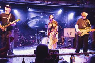 Agatha Is Dead on a small stage under a brick arch, singer in a mesh sweater at the mic, flanked by a guitarist in a cap and sunglasses and a bassist, drummer in the back. Purple-blue light.