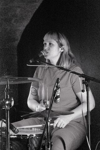 Black and white shot of Ellis Frawley singing from behind the drum kit, with cymbals and mic stands in the foreground.