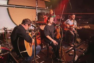 When The Robin Sings playing a seated set: singer holding a violin, acoustic guitarist on the left, bassist on the right, keys player behind them.