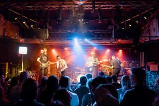 Chest playing to a packed crowd, two guitarists, a bassist and a singer hunched over the mic, drummer in the back. Red and blue stage lights.
